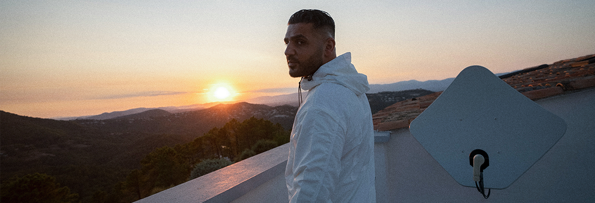 Es ist der Rapper Kurdo zu sehen, der auf einer Dachterasse steht, während im Hintergrund die Sonne untergeht.