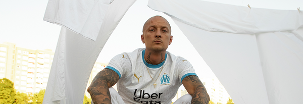 Es ist der Rapper Kurdo zu sehen, der auf einer Dachterasse steht, während im Hintergrund die Sonne untergeht.